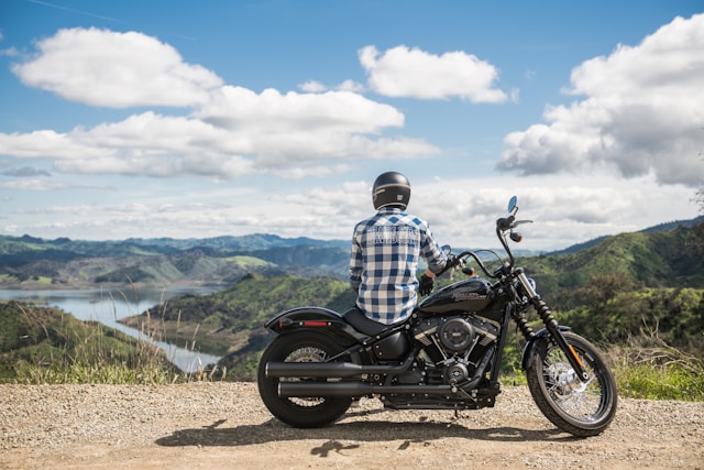 Man in blue and white checkered button down sitting on a motorcycle and looking out at a view of a river and mountains.