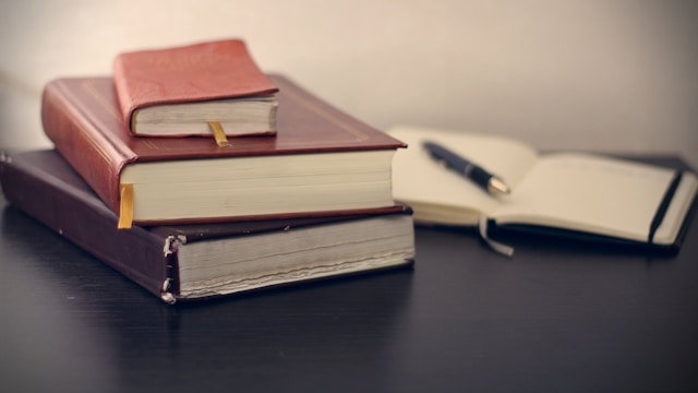 A stack of books next to an open notebook with a pen resting on top.