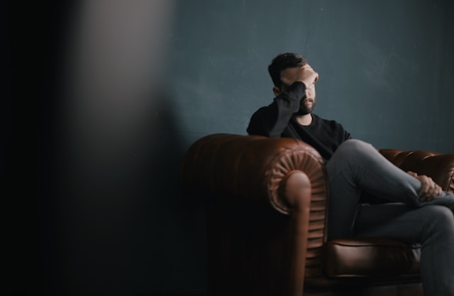 Man sitting on brown, leather couch with his hand on his forehead.